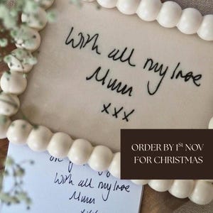 Puede incluir: Una placa rectangular de tela con texto bordado en negro en cursiva que dice With all my love Mum xxx. La placa está enmarcada por un borde de cuentas esféricas de color blanco roto, lisas y mate, dispuestas en un bucle continuo.