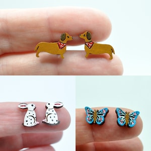 May include: A collection of wooden earrings featuring animal designs. The earrings include two dachshunds with red bandanas, two white rabbits with black spots, and two butterflies with blue and pink wings. The earrings are small and colorful.
