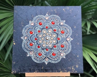 Könnte beinhalten: Quadratische schwarze Leinwand mit einem weißen und roten Mandala-Design. Das Mandala weist komplizierte Muster auf und ist mit roten und goldenen Verzierungen versehen. Die Leinwand wird auf einer kleinen Holzstaffelei präsentiert. Der Text "candlestrophic_designs" ist sichtbar.