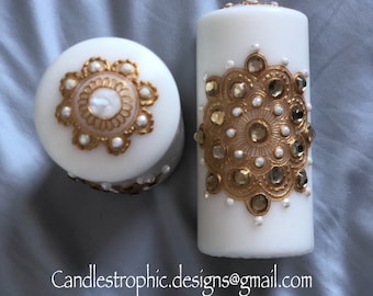 Könnte beinhalten: Zwei weiße Säulenkerzen mit goldenen und perlenartigen Verzierungen. Eine Kerze ist zylindrisch, die andere rund. Die Kerzen sind mit einem goldenen Filigranmuster und kleinen Perlen verziert. Der Text "Candlestrophic_designs" ist sichtbar.