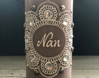 Könnte beinhalten: Eine mauvefarbene Säulenkerze mit einem weißen Blumenmuster und dem Schriftzug "Nan". Die Kerze ist mit klaren und grauen Kristallen verziert. Ein dekoratives Wohnaccessoire. Die Kerze ist etwa 15 cm hoch.