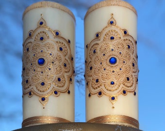 Könnte beinhalten: Zwei elfenbeinfarbene Säulenkerzen mit goldenen und blauen Verzierungen. Jede Kerze hat ein goldenes Filigranmuster mit blauen Akzenten und einem goldenen Band. Die Kerzen stehen auf einem goldfarbenen Sockel. Der Text "Candlestrophic designs" ist sichtbar.