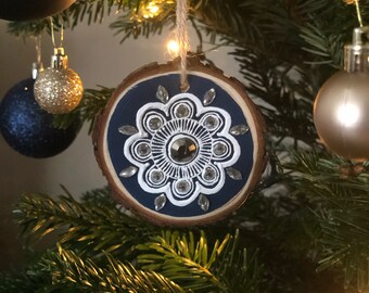 Könnte beinhalten: Ein Weihnachtsbaumschmuck aus Holzscheiben mit marineblauem Zentrum und weißem Blumenmuster. Der Schmuck ist mit silberfarbenen Strasssteinen und einem zentralen Edelstein verziert. Hängt an einem Weihnachtsbaum.