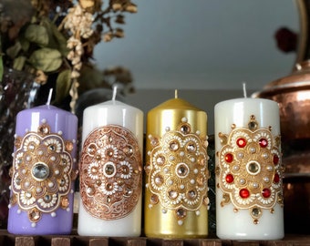 Könnte beinhalten: Vier Säulenkerzen mit aufwendigen, juwelenfarbenen Designs. Die Kerzen sind lavendelfarben, weiß, gold und weiß. Jede Kerze hat ein zentrales Design mit goldenen, weißen und Schmuckakzenten. Die Kerzen sind ein dekoratives Wohnaccessoire.