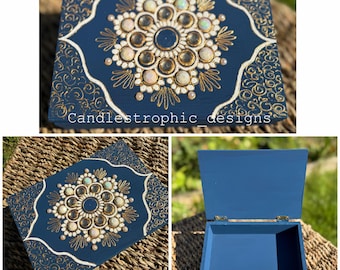 Könnte beinhalten: Eine marineblaue Holzbox mit dekorativem Design. Der Deckel hat ein zentrales Blumenmuster mit opalisierenden Steinen und goldenen Akzenten. Der Text "Candlestrophic designs" ist sichtbar.