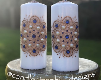 Könnte beinhalten: Zwei weiße Säulenkerzen mit aufwendigen goldenen und blauen Verzierungen. Das Design zeigt ein symmetrisches Muster aus goldenem Filigran, blauen Edelsteinen und Perlenakzenten. Die Kerzen werden auf einem dekorativen Ständer präsentiert.
