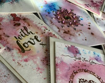 Könnte beinhalten: Eine Sammlung handgefertigter Grußkarten mit Aquarellhintergründen in Rosa-, Lila- und Blautönen. Einige Karten haben Verzierungen wie Strasssteine und Texte wie "With Love" und "Just For You".
