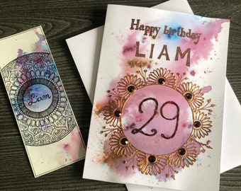 Könnte beinhalten: Geburtstagskarte mit dem goldenen Schriftzug "Happy birthday LIAM". Die Zahl "29" befindet sich in der Mitte eines floralen Designs. Ein passendes Lesezeichen mit dem Namen "Liam" ist ebenfalls vorhanden.