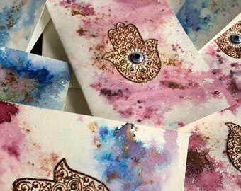 Könnte beinhalten: Aquarell-Grußkarten mit einem Hamsa-Hand-Design. Die Karten zeigen einen rosa, blauen und goldenen Aquarellhintergrund. Die Hamsa-Hand ist mit goldenem Glitzer umrandet und hat ein blaues Auge in der Mitte. Der Text "Candlestrophic_designs" ist sichtbar.