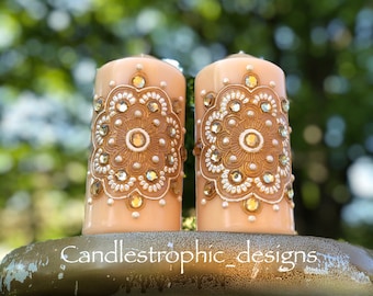 Könnte beinhalten: Zwei pfirsichfarbene Säulenkerzen mit aufwendigen Gold- und Kristallverzierungen. Die Kerzen weisen ein detailliertes Mandala-ähnliches Design auf. Der Text "Candlestrophic_designs" ist auf dem Sockel zu sehen. Die Kerzen stehen auf einem goldfarbenen dekorativen Ständer.
