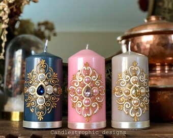 Könnte beinhalten: Drei dekorative Säulenkerzen in Blau, Rosa und Beige. Jede Kerze hat ein verziertes Golddesign mit Kunstperlen und einem zentralen Juwel. Die Kerzen haben ein silbernes Band am Boden und sind mit "Candlestrophic designs" beschriftet.