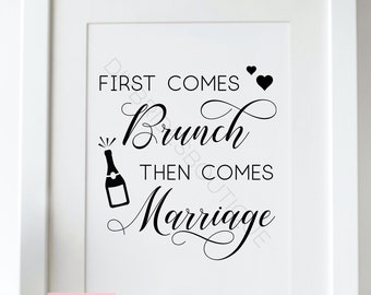 May include: A white framed print with the text "FIRST COMES Brunch THEN COMES Marriage" in black script. The image includes a champagne bottle illustration and two hearts. The print is on a white surface with greenery.