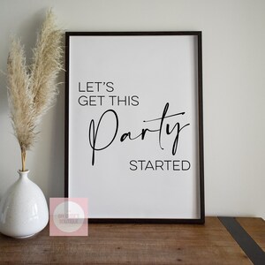 Könnte beinhalten: Ein schwarzer gerahmter Druck mit weißem Hintergrund und schwarzem Text, der "Let's Get This Party Started" lautet. Der Druck liegt auf einem Holztisch mit einer weißen Vase mit getrocknetem Pampasgras.