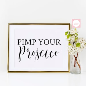 May include: A gold framed print with the text "PIMP YOUR Prosecco" in black calligraphy. The print is on a white background and is sitting on a white surface. There is a vase with white flowers to the right of the print.