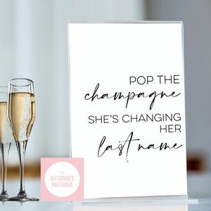 Könnte beinhalten: Ein weißes Schild mit schwarzem Kalligraphietext, der "Pop the champagne she's changing her last name" lautet. Im Vordergrund stehen zwei mit Champagner gefüllte Sektgläser.