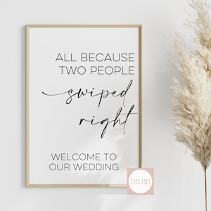 May include: A white sign with a gold frame, featuring the text "All because two people swiped right. Welcome to our wedding." The sign is leaning against a white wall with a bouquet of dried pampas grass in the background.