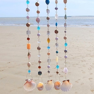 Puede incluir: Cinco hilos decorativos de cuentas colgantes con temática de playa. Cada hilo presenta una mezcla de cuentas coloridas de varias formas y tamaños, incluyendo transparentes, azules y marrones. Los hilos están terminados con una concha en la parte inferior, sobre un fondo de playa.