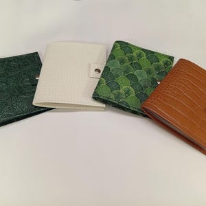 Puede incluir: Cuatro carteras cuadradas con cierres de presión dispuestas en fila. La primera es verde oscuro con un patrón floral en relieve, la segunda es blanco roto con un tejido texturizado, la tercera es verde con un patrón de abanico y la cuarta es marrón con textura de piel de cocodrilo.