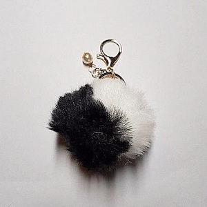 Könnte beinhalten: Ein schwarzer und weißer flauschiger Pompon-Schlüsselanhänger mit einem goldenen Schlüsselring und einem kleinen Perlenanhänger.