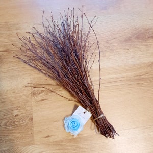 Può includere: Un fascio di ramoscelli marroni legati insieme con uno spago. Un piccolo fiore blu con un bordo di pizzo bianco è attaccato a un'etichetta bianca che dice "Baskets".