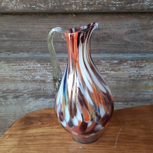 Könnte beinhalten: Eine farbenfrohe Glaskaraffe mit einem Wirbelmuster in Weiß-, Orange-, Braun- und Blautönen. Die Karaffe hat einen klaren Glasgriff.