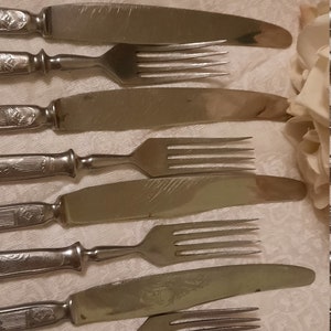 May include: A set of vintage silverware, including knives and forks, arranged on a white tablecloth. The silverware has ornate handles and a tarnished appearance, suggesting age and use. The forks have four tines.