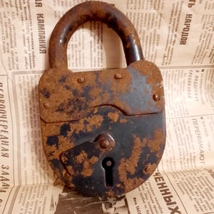 May include: An old, rusty padlock with a curved handle, displaying a dark brown, weathered appearance. The lock is positioned on a backdrop of aged newspaper print, emphasising its vintage style.
