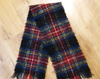 May include: A plaid scarf in a classic tartan pattern. The scarf features a black base with red, blue, yellow, and white intersecting lines. The ends of the scarf have a fringed trim. The scarf is made of a soft, woven material.