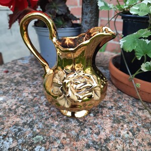 May include: A small, gold-coloured jug with a handle and a spout. The jug features a raised rose design on the front. The shiny, metallic finish reflects the surrounding light. A decorative item for display or serving.
