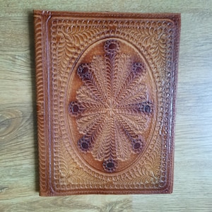 May include: A brown leather journal with an embossed floral design. The cover features an oval centerpiece with a detailed, symmetrical pattern and dark brown accents. The edges are also embossed with a repeating design.