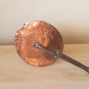 Può includere: Una padella di rame con un lungo manico in metallo scuro. La padella ha una superficie rotonda e martellata con un ricco colore marrone-rossastro. Il manico è fissato con rivetti e ha un anello all'estremità.