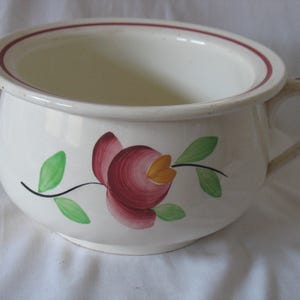 Peut inclure: Pot de chambre en céramique blanc cassé avec une poignée et un motif floral. Le pot a un large bord avec une rayure marron et une fleur peinte avec des pétales marron, des feuilles vertes et un centre jaune.
