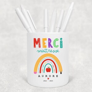 Peut inclure: Un pot à crayons en céramique blanche avec les mots "MERCI maîtresse" en lettres colorées, un motif arc-en-ciel et le nom "AURORE" en dessous. Le pot est rempli de stylos blancs.
