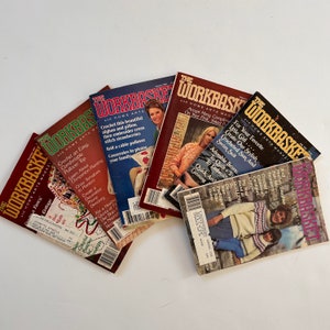 May include: A collection of vintage "Workbasket" magazines, featuring colorful covers with images of crafts, knitting, and home arts. The magazines are stacked and fanned out, showcasing various issues from different months.