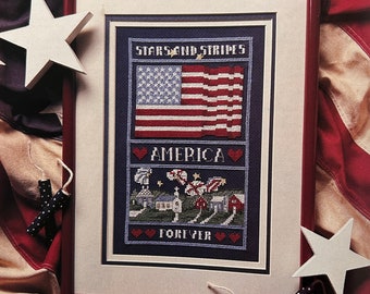 May include: A framed cross-stitch design featuring an American flag with the text "Stars and Stripes" above it, a small town scene with the text "America" above it, and the text "Forever" below it. The frame is red and the background of the cross-stitch is dark blue.