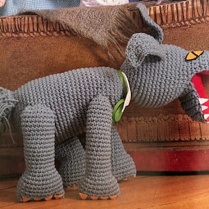 May include: A gray crocheted wolf with a red open mouth and white teeth. The wolf has a green bow tie and orange feet.