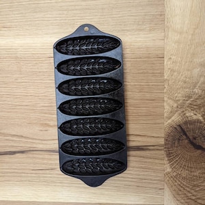 May include: Black cast iron loaf pan with six individual molds shaped like wheat stalks. The pan has a handle on the top.