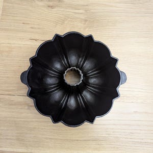 May include: A black cast iron bundt pan with a fluted design. The pan has a central tube and two handles on the sides. The pan is sitting on a light brown wooden surface.