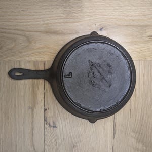May include: A black cast iron skillet with a single handle. The skillet has a smooth, black surface and a letter "L" embossed on the bottom.