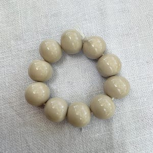 Könnte beinhalten: Eine kreisförmige Anordnung aus elf glatten, glänzenden, beigefarbenen, kugelförmigen Perlen, die auf einer dünnen weißen Schnur aufgereiht sind. Jede Perle hat eine reflektierende Oberfläche und eine gleichmäßige, runde Form, die eine einfache Schleife bildet.
