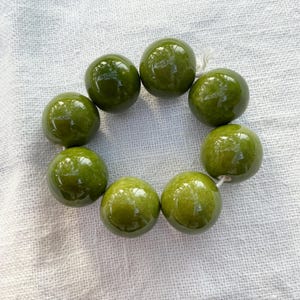 Könnte beinhalten: Eine kreisförmige Anordnung aus acht glatten, glänzenden, olivgrünen, kugelförmigen Perlen, die auf einer dünnen weißen Schnur aufgereiht sind. Jede Perle hat eine reflektierende Oberfläche mit subtilen Variationen im tiefen Grün für ein einheitliches Bild.
