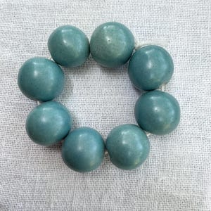Può includere: Una disposizione circolare di otto perline sferiche lisce, opache e di colore blu-verde, infilate su un sottile cordino bianco. Le perline hanno una forma rotonda coerente e una finitura blu-verde solida e uniforme, creando un anello semplice.