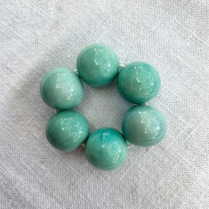 Può includere: Una disposizione circolare di sei perline in ceramica rotonde, lucide e di colore turchese. Ogni perlina presenta una sottile e fine finitura smaltata a crepa sulla superficie. Le perline sono infilate su un sottile cordino bianco.