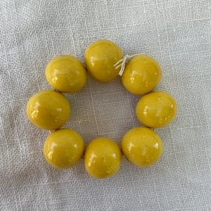 Può includere: Un braccialetto circolare composto da otto perle sferiche color giallo senape, lisce e lucide. Le perle sono infilate su un cordino sottile di colore chiaro, visibile come un piccolo nodo tra due perle, creando un anello continuo.