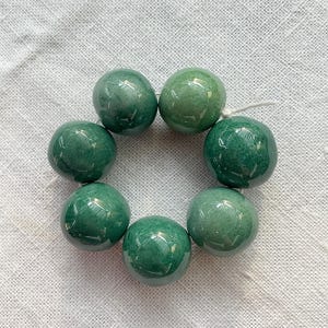 Pode incluir: Um arranjo circular de sete contas verdes esféricas e lisas enfiadas num fio branco fino. Cada conta apresenta um acabamento polido e brilhante com variações subtis na cor verde floresta e na textura interna do material.
