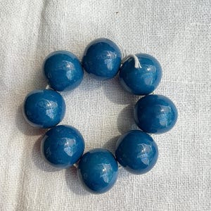Pode incluir: Uma pulseira circular composta por oito contas esféricas grandes, lisas e de um tom azul-petróleo profundo. As contas estão enfiadas num fio fino de cor clara, visível no topo do arranjo, formando um círculo contínuo.