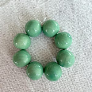 Può includere: Un braccialetto con otto grandi perline rotonde verde chiaro. Le perline sono infilate con un filo bianco, formando una forma circolare. Le perline hanno una finitura lucida e riflettono la luce. Il braccialetto è esposto su una superficie bianca strutturata.