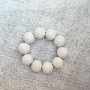 Könnte beinhalten: Eine kreisförmige Anordnung von elf weißen, glänzenden, runden Perlen. Die Perlen sind auf einer dünnen, weißen Schnur aufgereiht. Die Perlen liegen auf einem strukturierten, cremefarbenen Stoff.