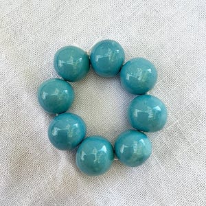 Op de afbeelding: Een cirkelvormige opstelling van acht gladde, glanzende, blauwgroene bolvormige kralen die aan een dun wit koord zijn geregen. Elke kraal heeft een reflecterend oppervlak met subtiele lichtinval, wat zorgt voor een uniforme uitstraling.