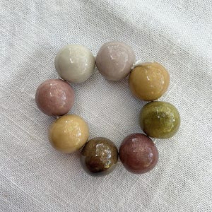 Könnte beinhalten: Eine kreisförmige Anordnung aus acht glatten, glänzenden, kugelförmigen Keramikperlen in verschiedenen Erdtönen wie Creme, Mauve, Senfgelb, Olivgrün und Dunkelbraun, die auf einer dünnen weißen Schnur zu einer durchgehenden Schlaufe aufgereiht sind.